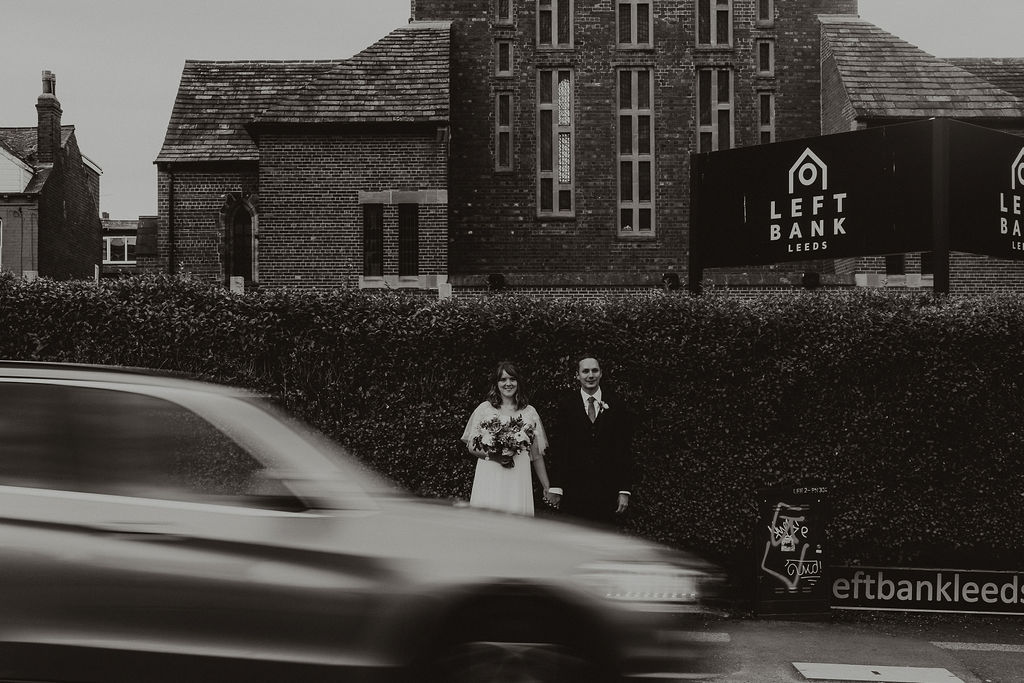 Bride and groom stood outside Left Bank Leeds with a blurry car speeding past in black and white